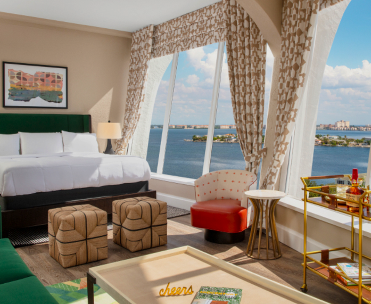 A stylish hotel room with a scenic window view, a bed with white linens, and modern furnishings, including ottomans and a bar cart.