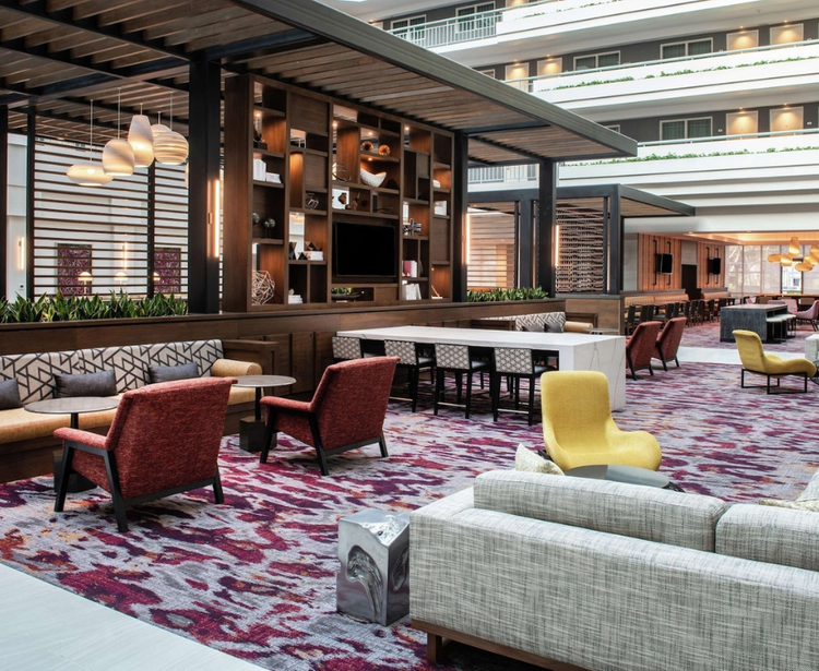 A modern hotel lobby with red and yellow seating, a patterned carpet, wooden shelves, and decorative accents, creating a welcoming atmosphere.